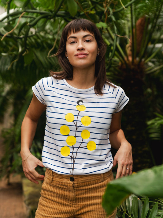 Weiß‑blau gestreiftes Damen‑T‑Shirt im Cap‑Sleeve‑Stil, leicht tailliert, mit weitem Rundhalsausschnitt. Aus weichem, GOTS‑zertifizierter Bio‑Baumwolle, nachhaltig und fair produziert. Auf der Vorderseite befindet sich ein Print: eine gelbe Blume, auf der sich ein kleines Mädchen ausruht. Das Shirt vermittelt ein entspanntes Urlaubs‑Feeling