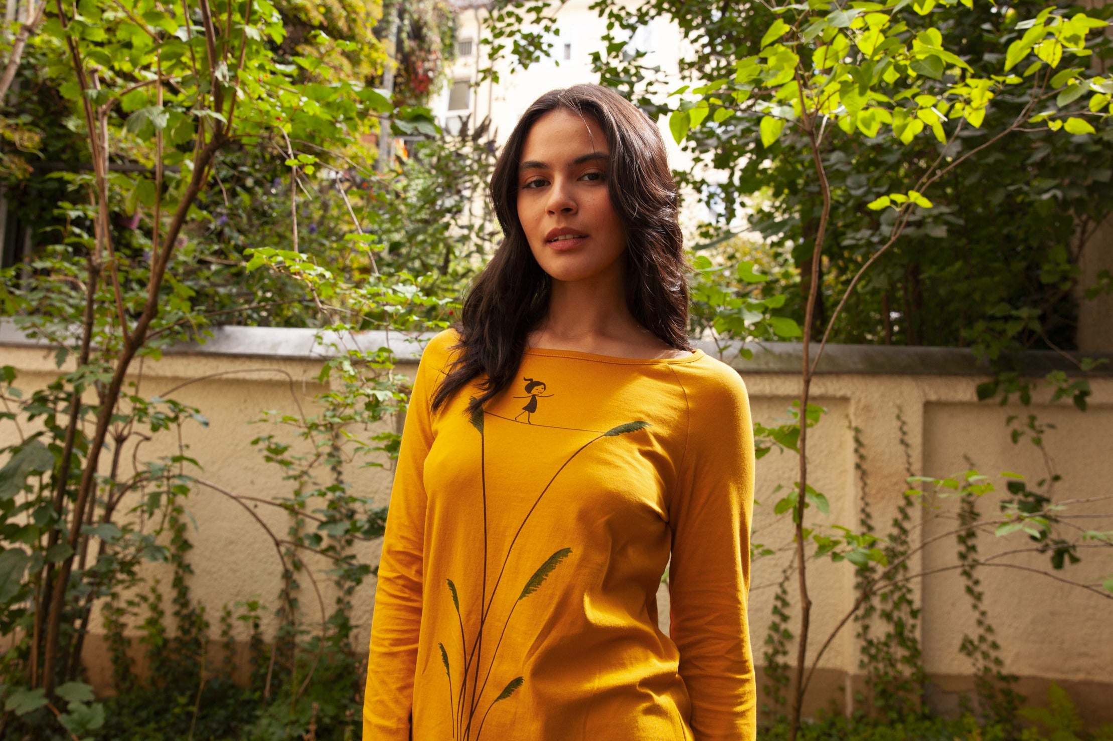 Model steht im begrünten Innenhof und trägt ein FellHerz Longsleeve mit dem Motiv Seiltanz in goldgelb