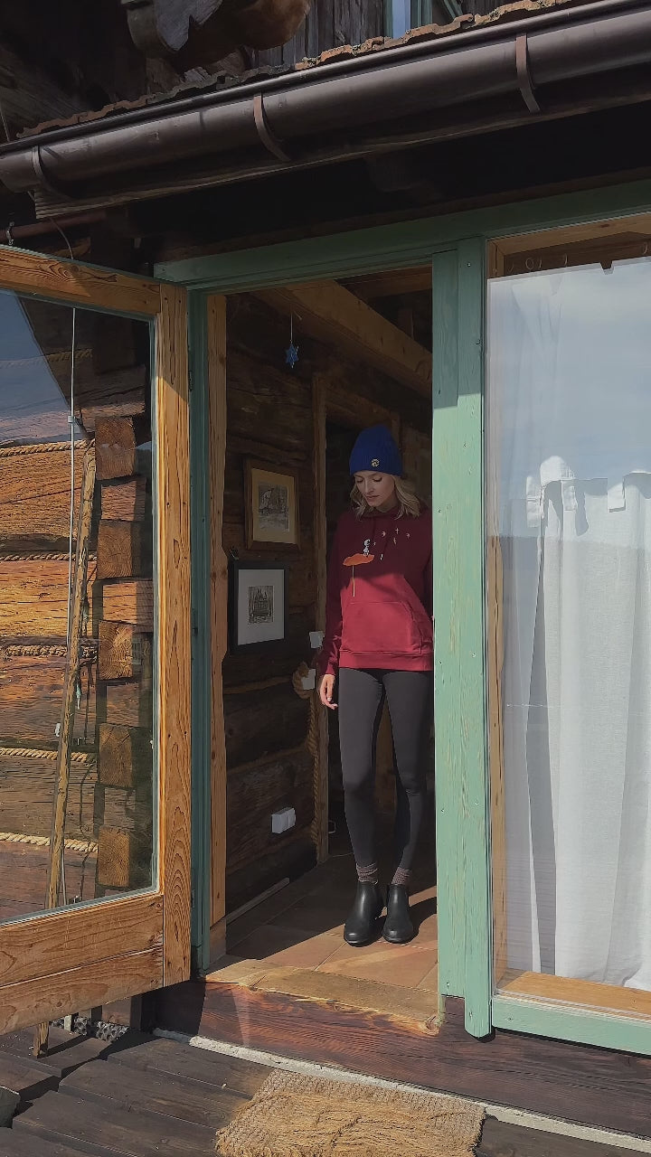 Video mit Model, es trägt einen dunkelroten damen kapuzen-pullover aus Bio-Baumwolle, fair und vegan produziert. Raglan-Schnitt mit Grafik-Print auf der Vorderseite über der kängurutasche: Ein Mädchen steht auf einer orangen Blume und es fliegen Vögel davon.