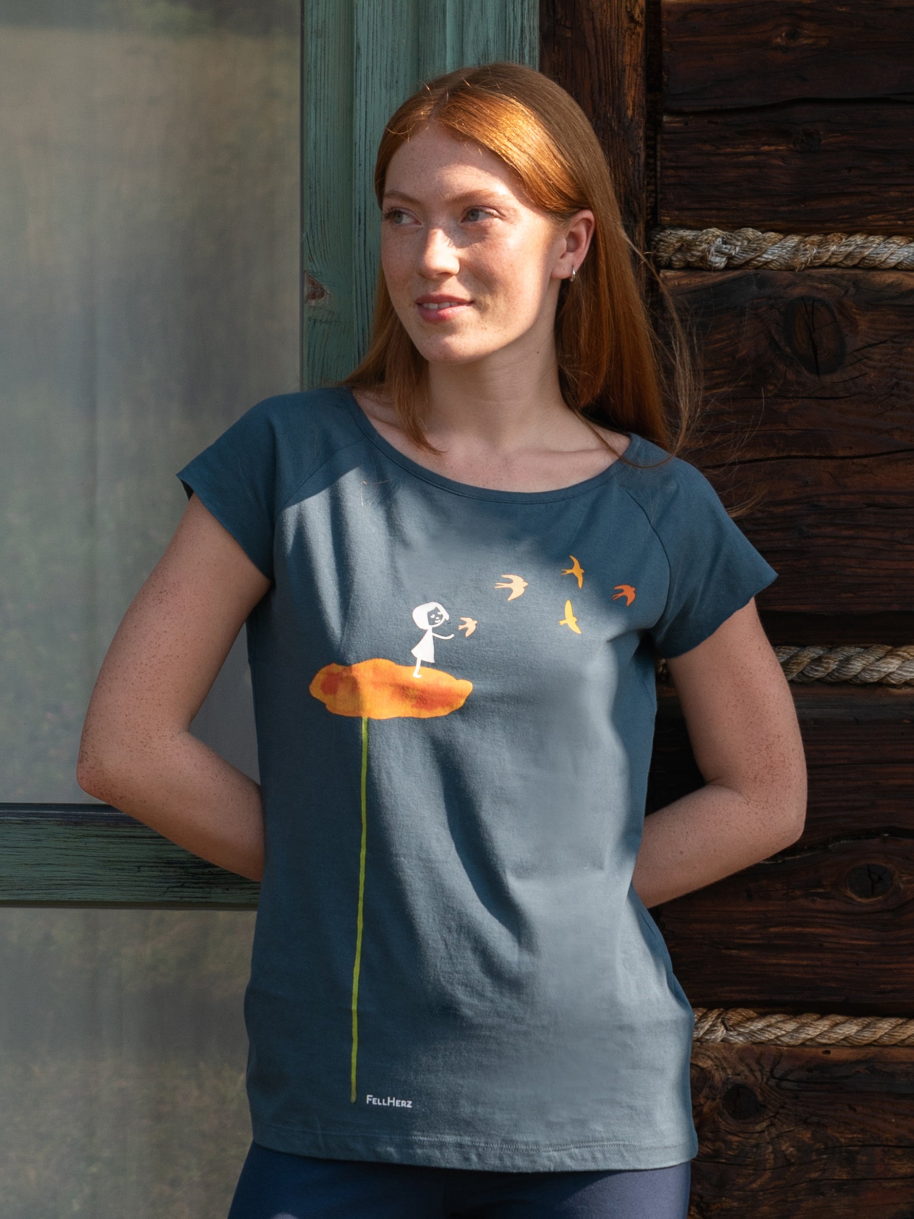 Mittel-blaues Damen T-Shirt aus Bio-Baumwolle, fair und vegan produziert. Raglan-Shirt mit Grafik-Print auf der Vorderseite: Ein Mädchen steht auf einer orangen Blume und es fliegen Vögel davon.