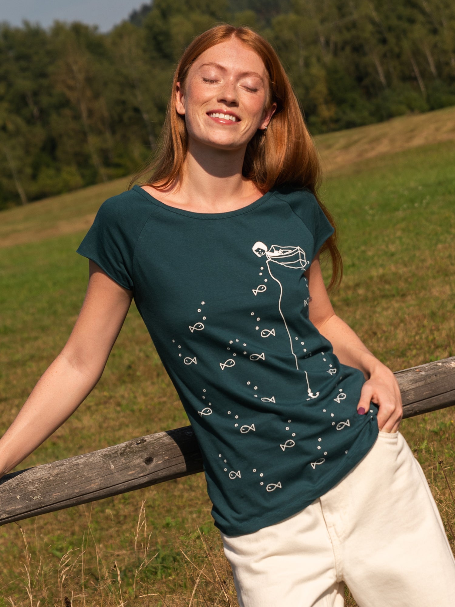 Ein Model trägt ein Damen T‑Shirt deep teal aus 100 % Bio‑Baumwolle mit kurzen Cap‑Sleeves, maritimem Anker‑Siebdruck vorne und kleinem Logo‑Print auf dem Rücken – nachhaltig, fair produziert und lässig geschnitten