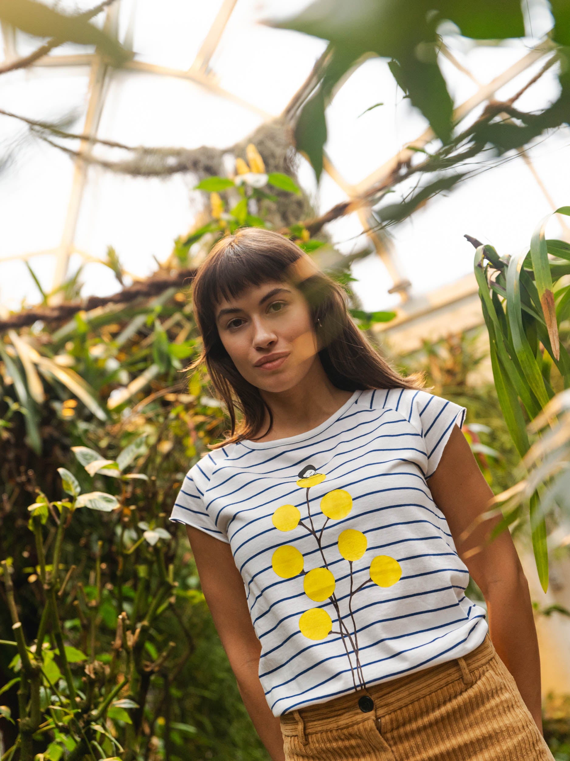 Weiß‑blau gestreiftes Damen‑T‑Shirt im Cap‑Sleeve‑Stil, leicht tailliert, mit weitem Rundhalsausschnitt. Aus weichem, GOTS‑zertifizierter Bio‑Baumwolle, nachhaltig und fair produziert. Auf der Vorderseite befindet sich ein Print: eine gelbe Blume, auf der sich ein kleines Mädchen ausruht. Das Shirt vermittelt ein entspanntes Urlaubs‑Feeling