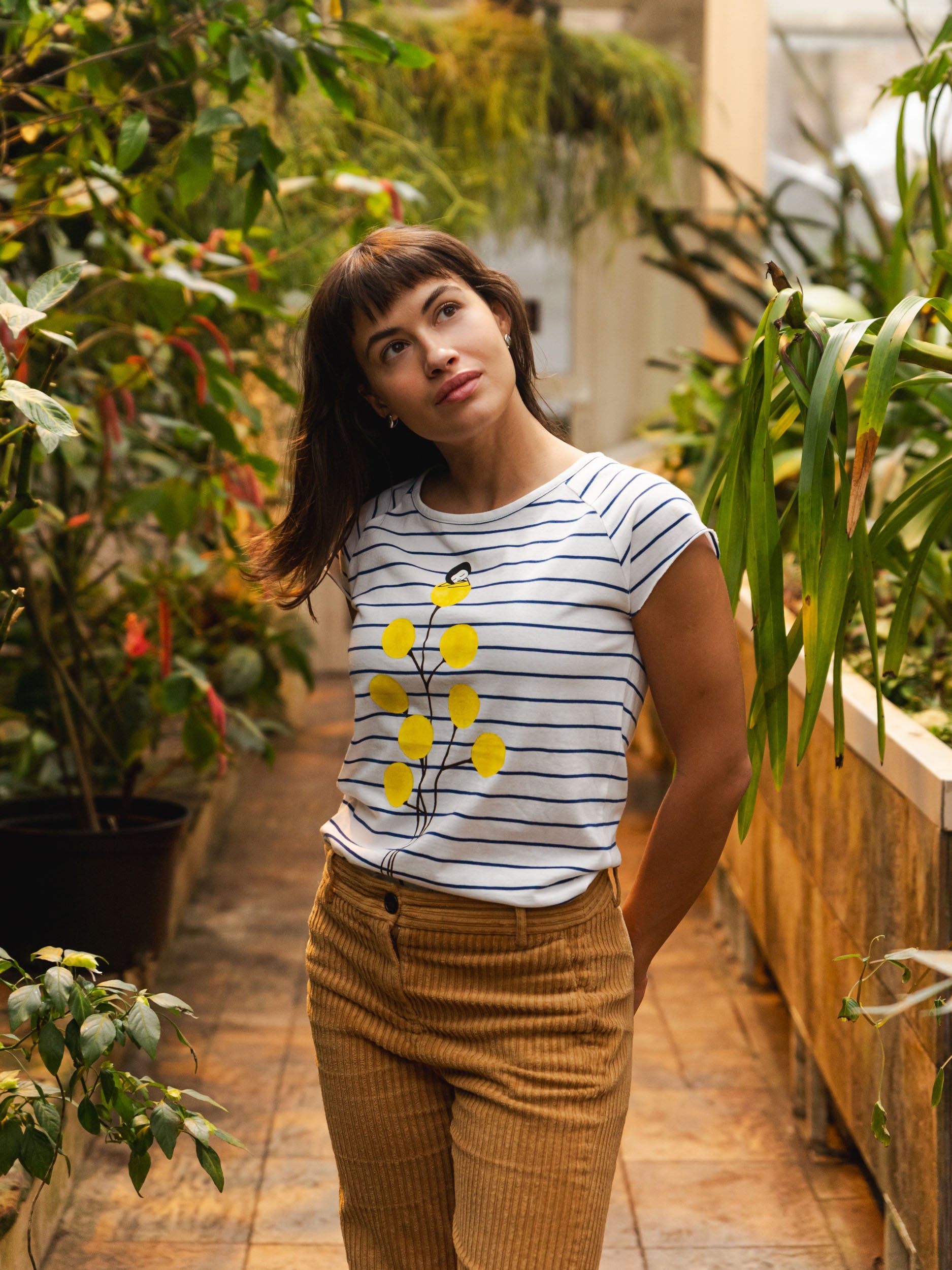 Weiß‑blau gestreiftes Damen‑T‑Shirt im Cap‑Sleeve‑Stil, leicht tailliert, mit weitem Rundhalsausschnitt. Aus weichem, GOTS‑zertifizierter Bio‑Baumwolle, nachhaltig und fair produziert. Auf der Vorderseite befindet sich ein Print: eine gelbe Blume, auf der sich ein kleines Mädchen ausruht. Das Shirt vermittelt ein entspanntes Urlaubs‑Feeling