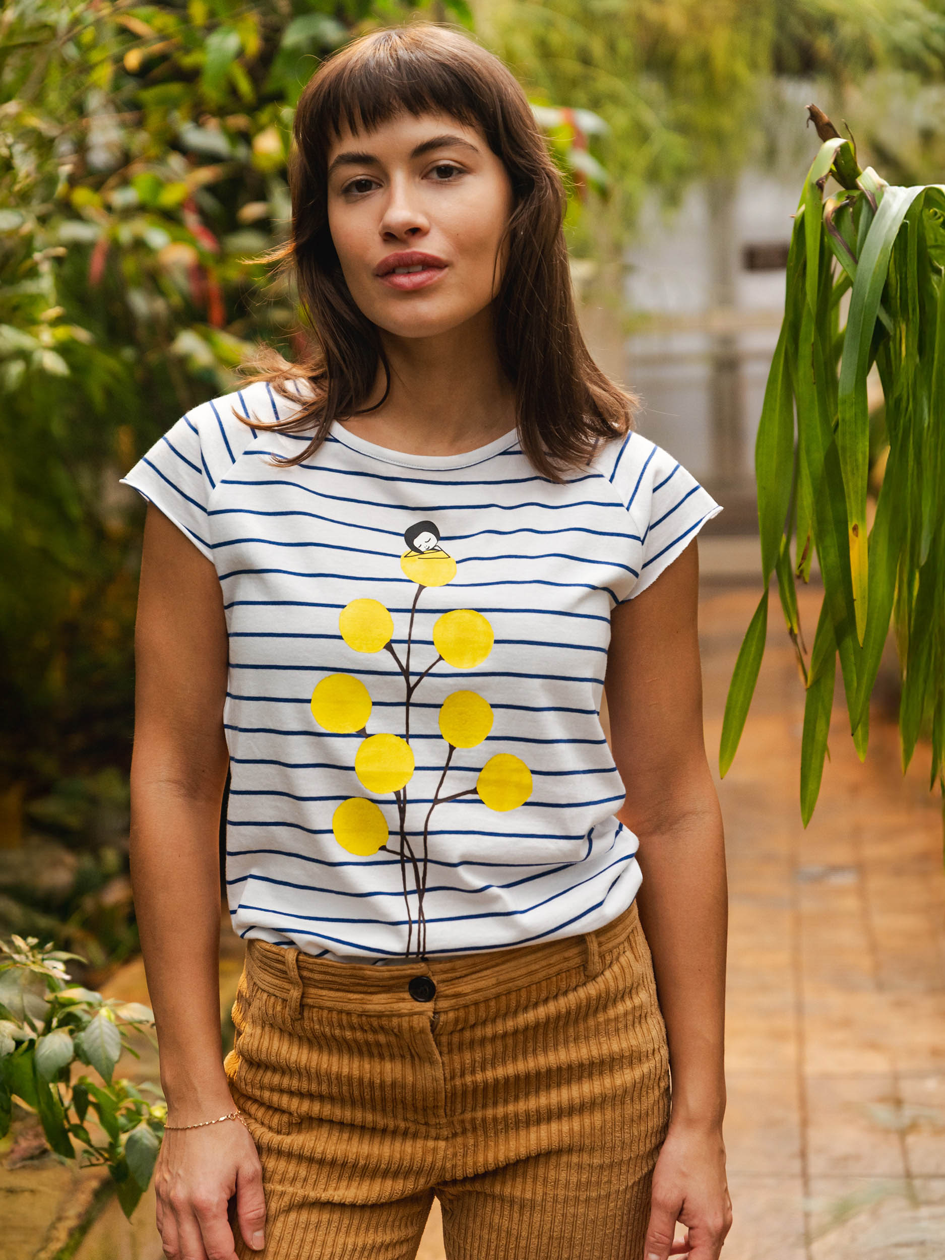 Weiß‑blau gestreiftes Damen‑T‑Shirt im Cap‑Sleeve‑Stil, leicht tailliert, mit weitem Rundhalsausschnitt. Aus weichem, GOTS‑zertifizierter Bio‑Baumwolle, nachhaltig und fair produziert. Auf der Vorderseite befindet sich ein Print: eine gelbe Blume, auf der sich ein kleines Mädchen ausruht. Das Shirt vermittelt ein entspanntes Urlaubs‑Feeling