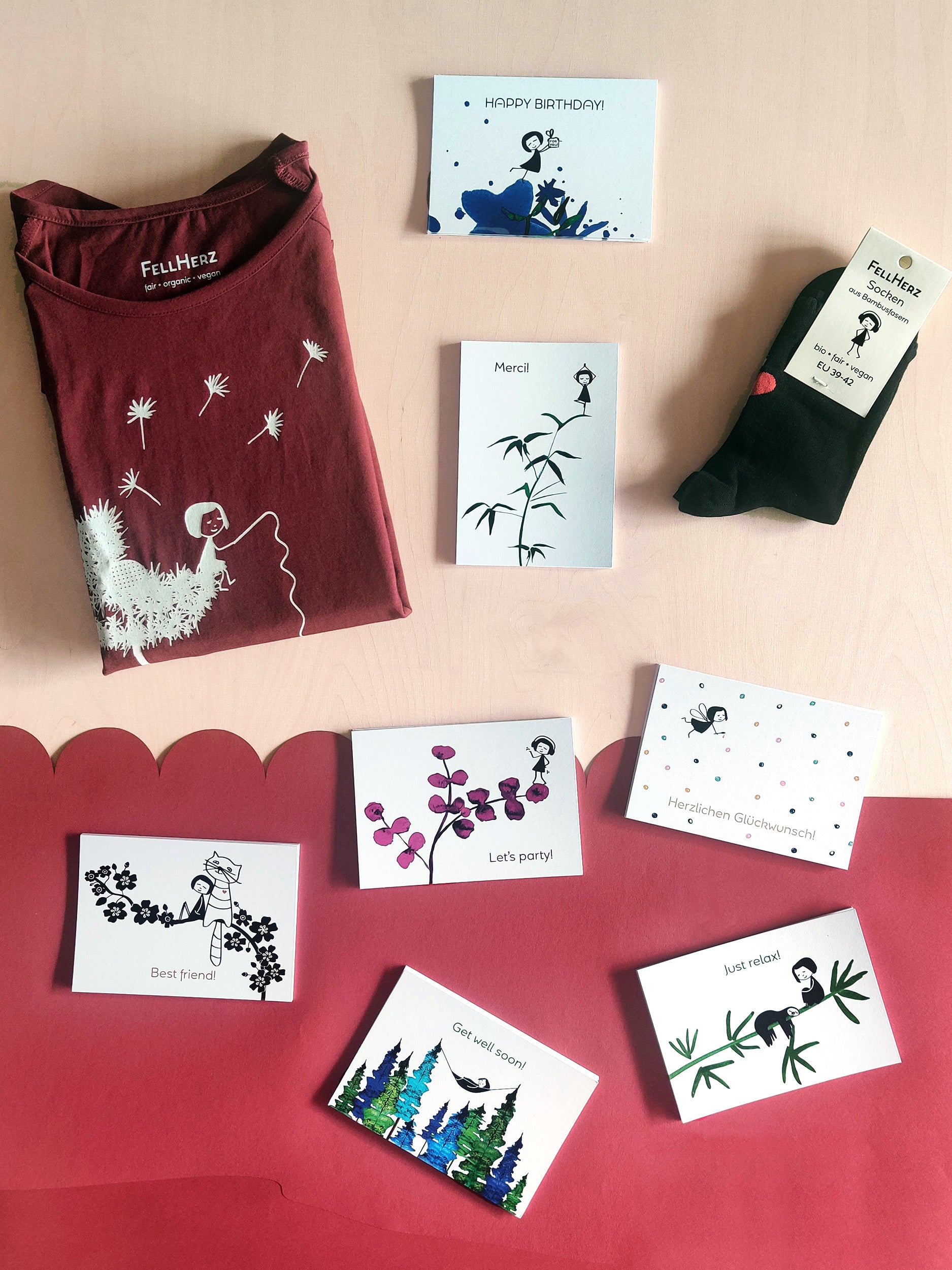 Ein Geschenk-Paket bestehend aus einem rubinrotem Bio-Baumwolle T-Shirt mit weißem Pusteblume Siebdruck, 7 unterschiedlichen Gruß-Postkarten und einem Paar schwarze Socken aus Bambusviskose mit kleinem roten Herz auf der Rückseite und Muschelsaum.