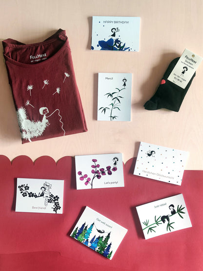 Ein Geschenk-Paket bestehend aus einem rubinrotem Bio-Baumwolle T-Shirt mit weißem Pusteblume Siebdruck, 7 unterschiedlichen Gruß-Postkarten und einem Paar schwarze Socken aus Bambusviskose mit kleinem roten Herz auf der Rückseite und Muschelsaum.