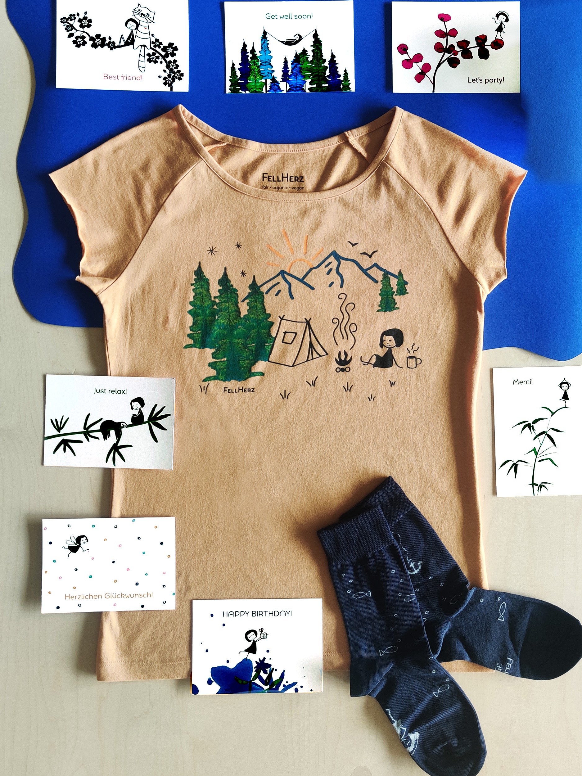 Ein Geschenk-Paket bestehend aus einem sandfarbenen Bio-Baumwolle T-Shirt mit einem Camping Druck (Zelt, Berge, Bäume und ein kleines Mädchen, das am Lagerfeuer sitzt, 7 unterschiedlichen Grußkarten und einem Paar dunkelblaue Socken mit weißen Fischen.