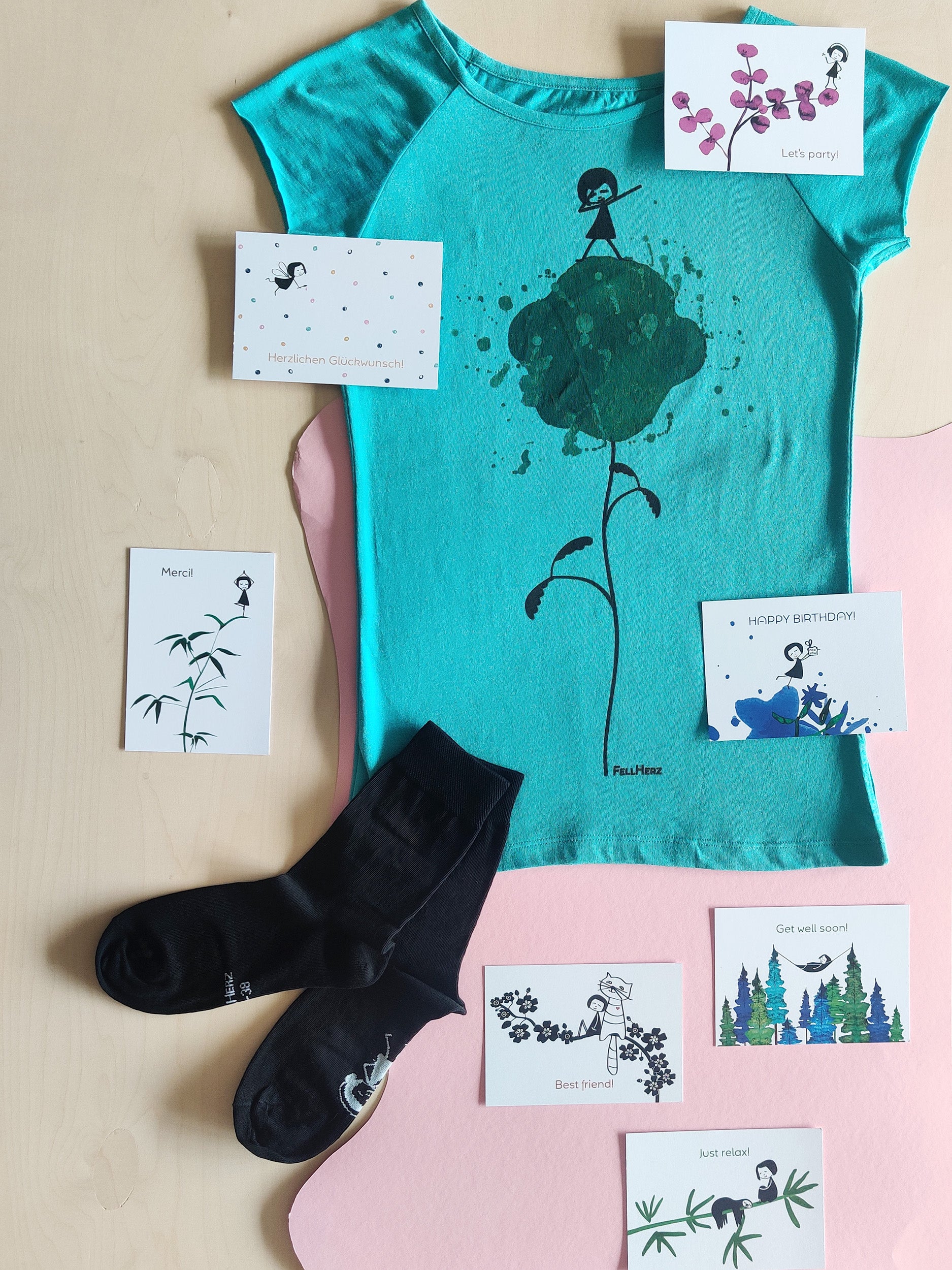 Ein Geschenk-Paket bestehend aus einem hellblauem Bio-Baumwolle T-Shirt mit einer Blumen, auf der ein kleines Mädchen tanzt , 7 unterschiedlichen Grußkarten und einem Paar schwarze Socken mit einem weißen Mädchen auf der Fußsohle.