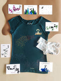 Ein Geschenk-Paket bestehend aus einem petrol Bio-Baumwolle Ommm T-Shirt mit buntem Regenbogen und einem Mädchen auf einem fliegenden Teppich, 7 unterschiedlichen Gruß-Postkarten und einem Paar weiße Bambus-Socken mit bunten Konfettitupfen und Muschelsaum.