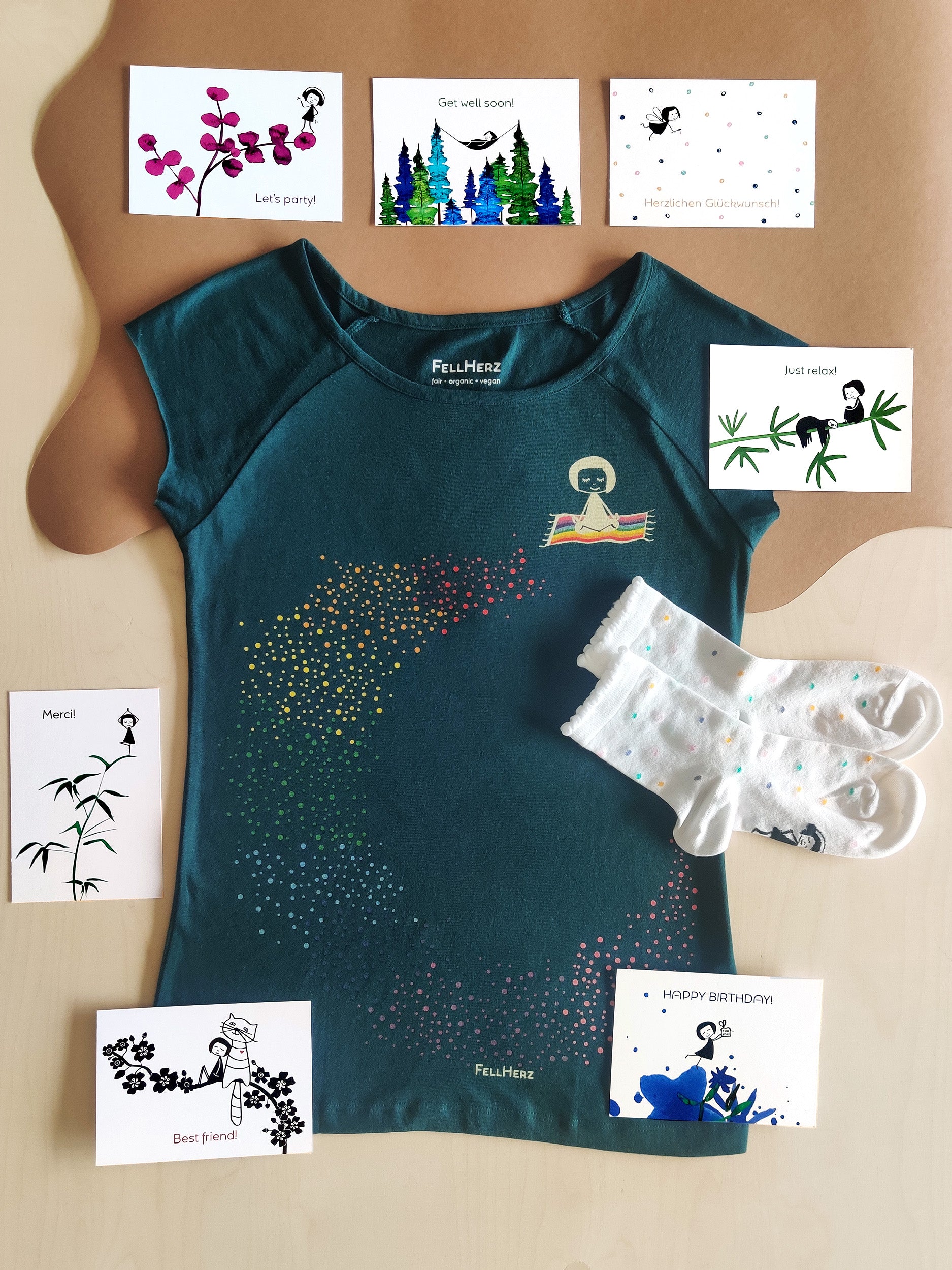 Ein Geschenk-Paket bestehend aus einem petrol Bio-Baumwolle Ommm T-Shirt mit buntem Regenbogen und einem Mädchen auf einem fliegenden Teppich, 7 unterschiedlichen Gruß-Postkarten und einem Paar weiße Bambus-Socken mit bunten Konfettitupfen und Muschelsaum.