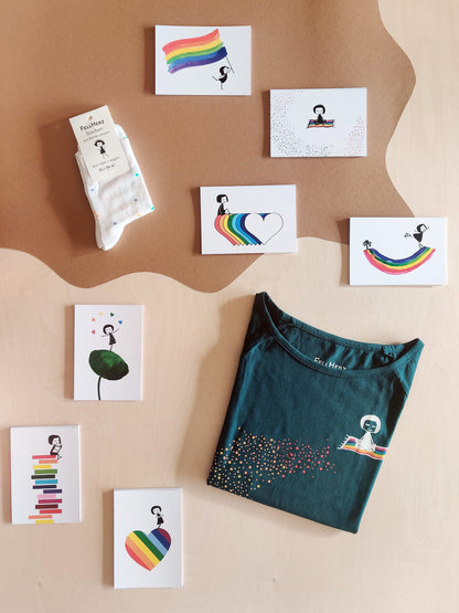 Ein Geschenk-Paket bestehend aus einem petrol Bio-Baumwolle Ommm T-Shirt mit buntem Regenbogen und einem Mädchen auf einem fliegenden Teppich, 7 unterschiedlichen Rainbow Postkarten und einem Paar weiße Bambus-Socken mit bunten Konfettitupfen und Muschelsaum.