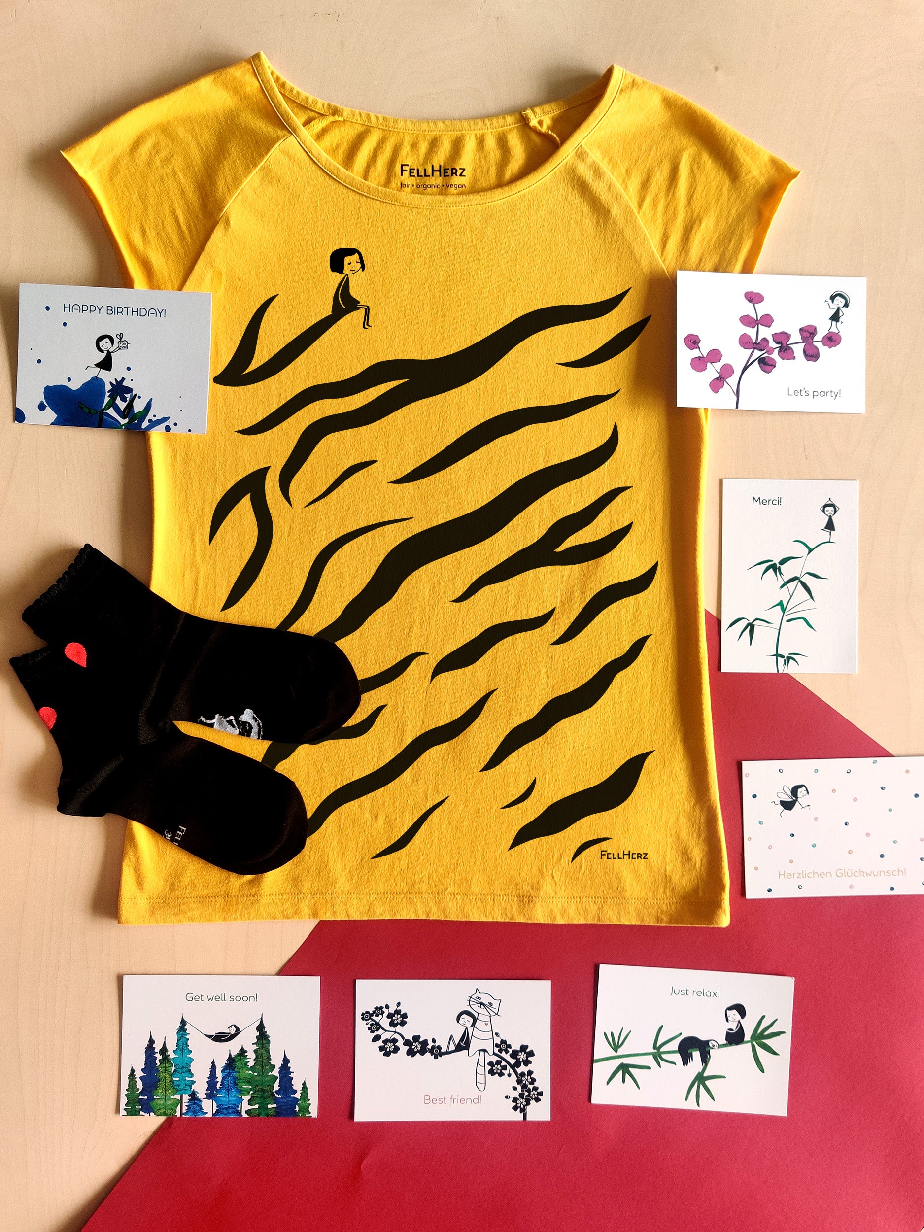 Ein Geschenk-Paket bestehend aus einem knallgelben Bio-Baumwolle T-Shirt mit schwarzem Siebdruck, ein Tigermuster Print mit kleinem sitzenden Mädchen. Dazu 7 unterschiedlichen Grußkarten und ein Paar schwarze Bambus-Socken mit einem kleinen roten Herz auf der Rückseite.