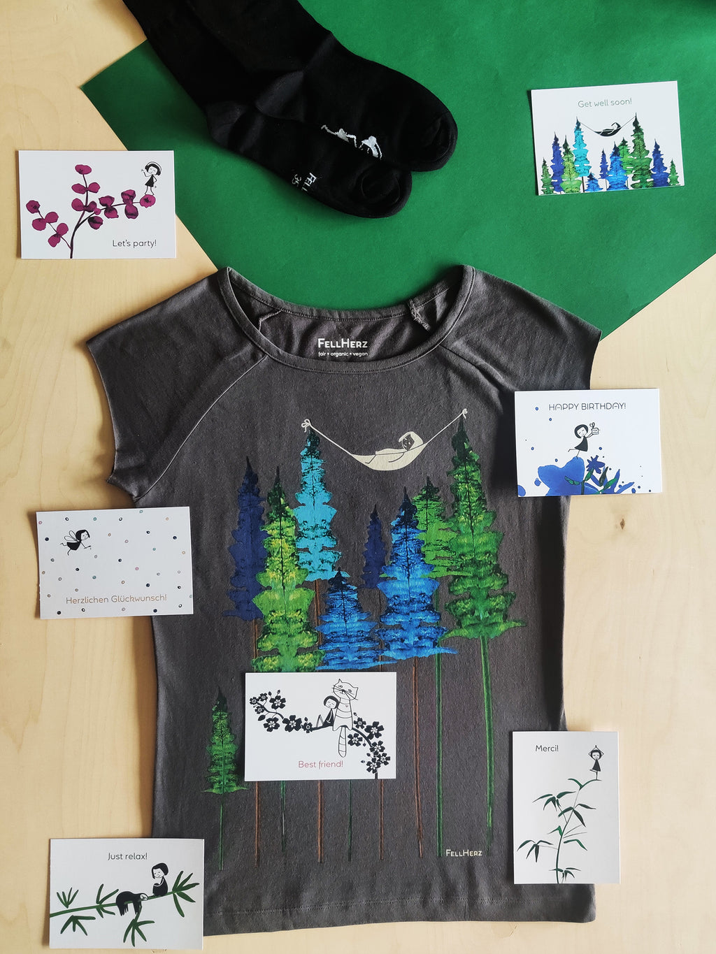 Ein Geschenk-Paket bestehend aus einem grauen Bio-Baumwolle T-Shirt mit einem Mädchen, das in einer Hängematte zwischen Bäumen liegt, 7 unterschiedlichen Grußkarten und einem Paar schwarze Socken mit einem kleinen Mädchenmotiv auf der Sohle.