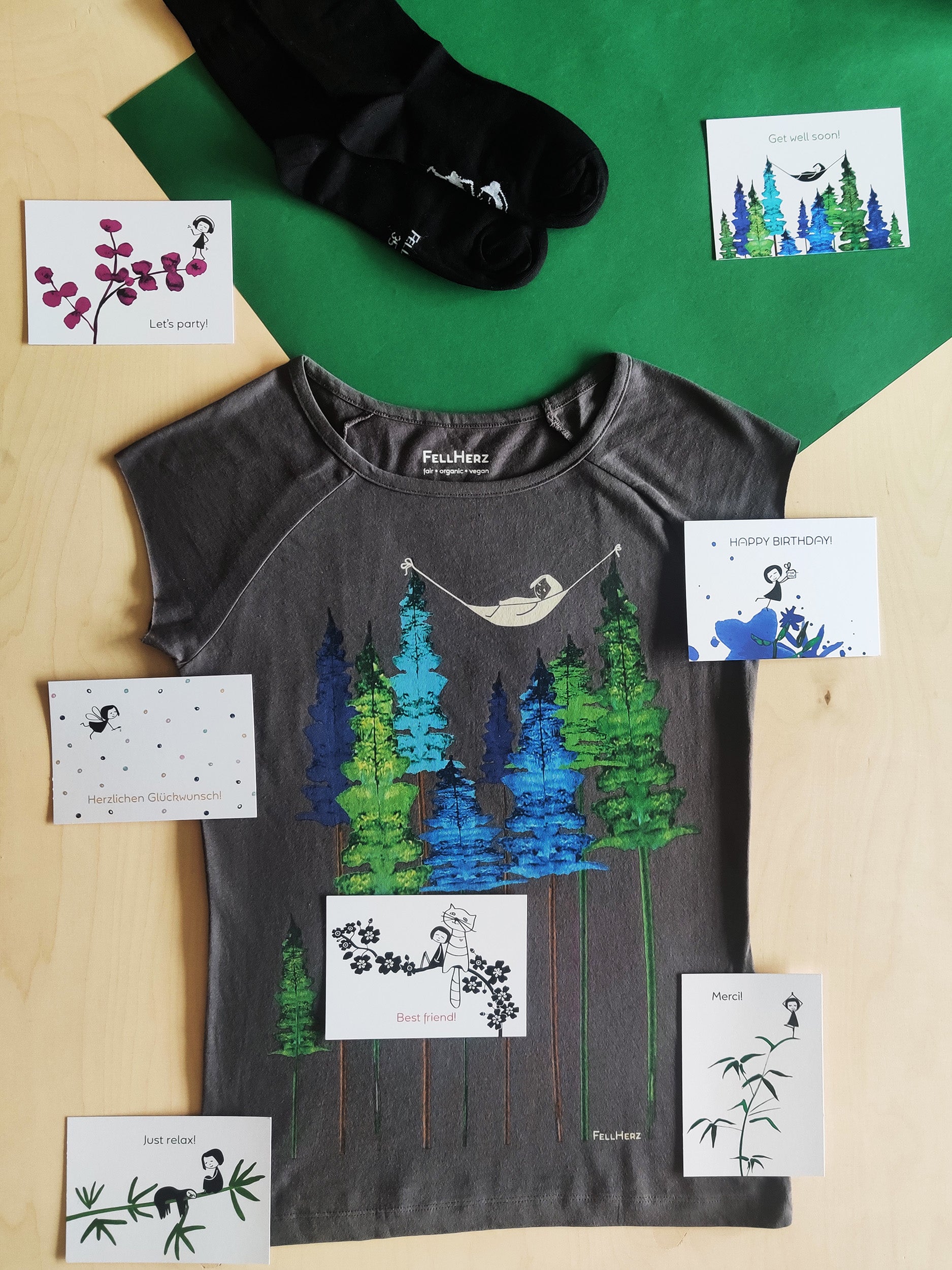 Ein Geschenk-Paket bestehend aus einem grauen Bio-Baumwolle T-Shirt mit einem Mädchen, das in einer Hängematte zwischen Bäumen liegt, 7 unterschiedlichen Grußkarten und einem Paar schwarze Socken mit einem kleinen Mädchenmotiv auf der Sohle.
