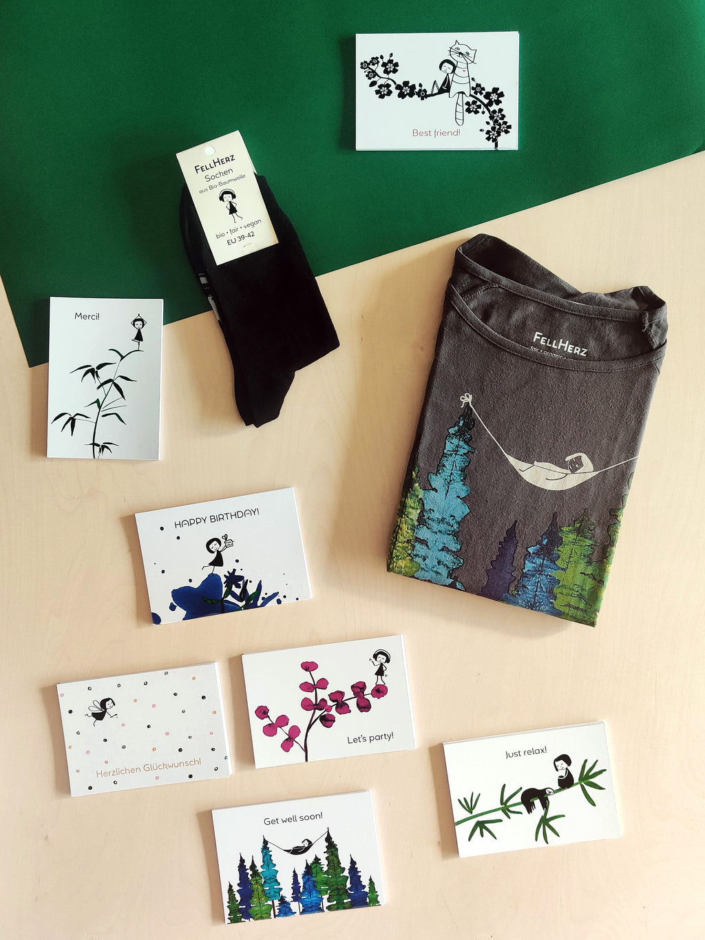 Ein Geschenk-Paket bestehend aus einem grauen Bio-Baumwolle T-Shirt mit einem Mädchen, das in einer Hängematte zwischen Bäumen liegt, 7 unterschiedlichen Grußkarten und einem Paar schwarze Socken mit einem kleinen Mädchenmotiv auf der Sohle.