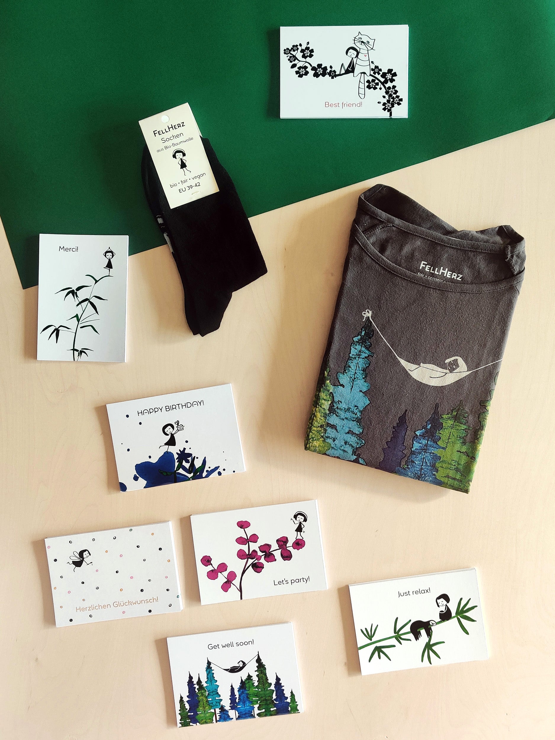 Ein Geschenk-Paket bestehend aus einem grauen Bio-Baumwolle T-Shirt mit einem Mädchen, das in einer Hängematte zwischen Bäumen liegt, 7 unterschiedlichen Grußkarten und einem Paar schwarze Socken mit einem kleinen Mädchenmotiv auf der Sohle.