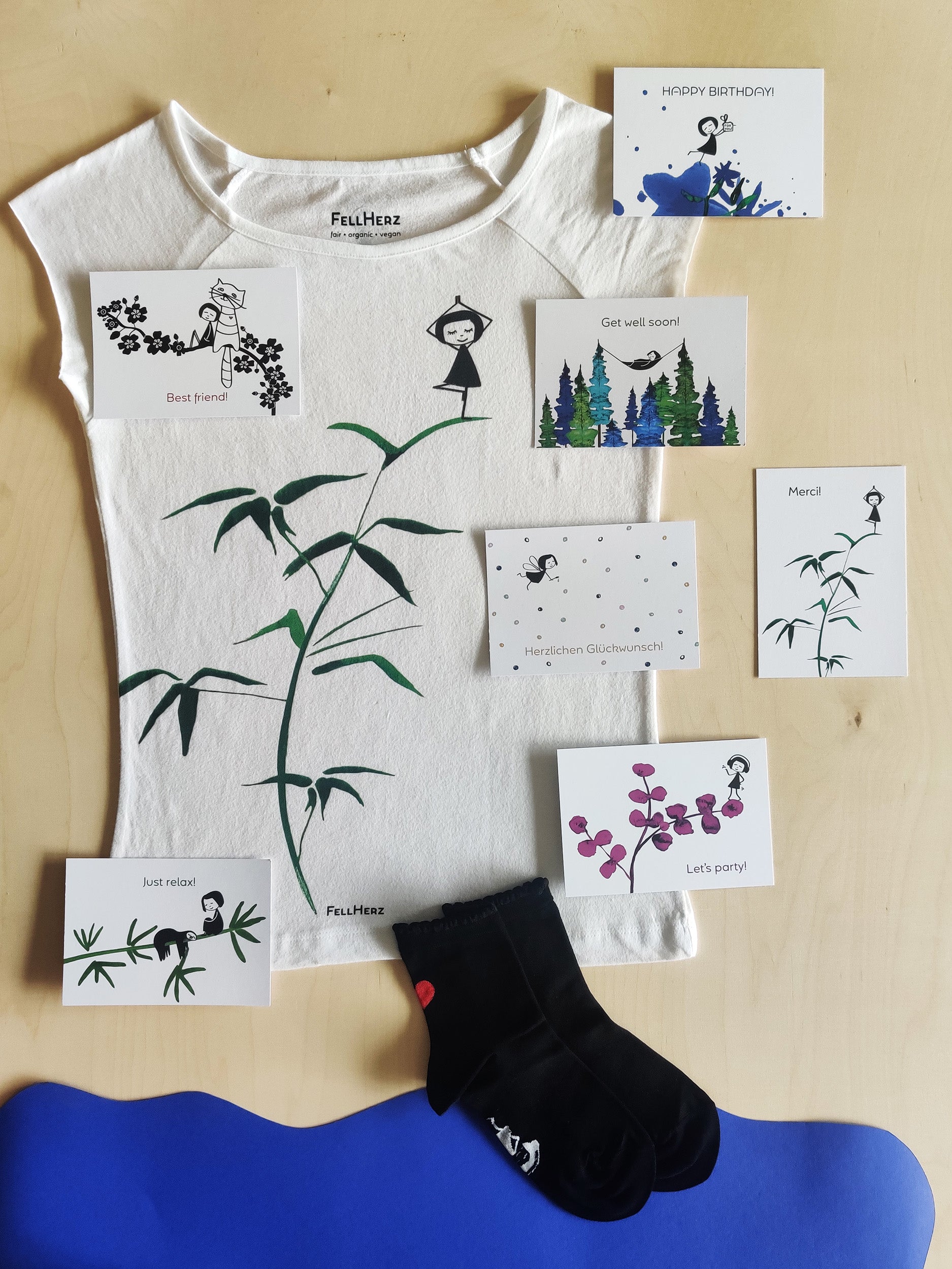 Ein Geschenk-Paket bestehend aus einem weißen Bio-Baumwolle T-Shirt mit bunten Konfettitfen und einem kleinen geflügelten Mädchen, 7 unterschiedlichen Gruß-Postkarten und einem Paar weiße Socken mit bunten Konfettitupfen und Muschelsaum.
