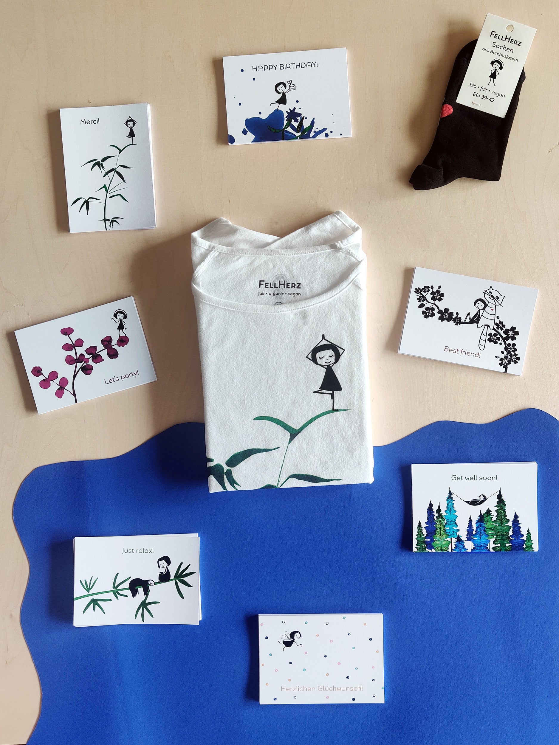 Ein Geschenk-Paket bestehend aus einem weißem Bio-Baumwolle T-Shirt mit einem Mädchen in Yogapose, das auf einem grünen Bambuszweig steht, 7 unterschiedlichen Grußkarten und einem Paar schwarze Socken mit Muschelsaum und roten Herz auf der Rückseite.