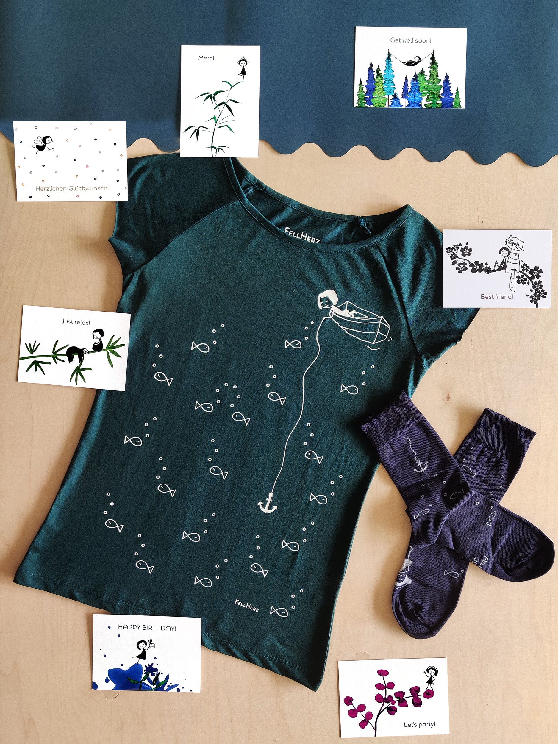 Ein Geschenk-Paket bestehend aus einem petrolfarbenen Bio-Baumwolle T-Shirt mit dem Motiv Ankermädchen, 7 unterschiedlichen Grußkarten und einem Paar blaue Socken mit weißem Anker und Fischmotiv.