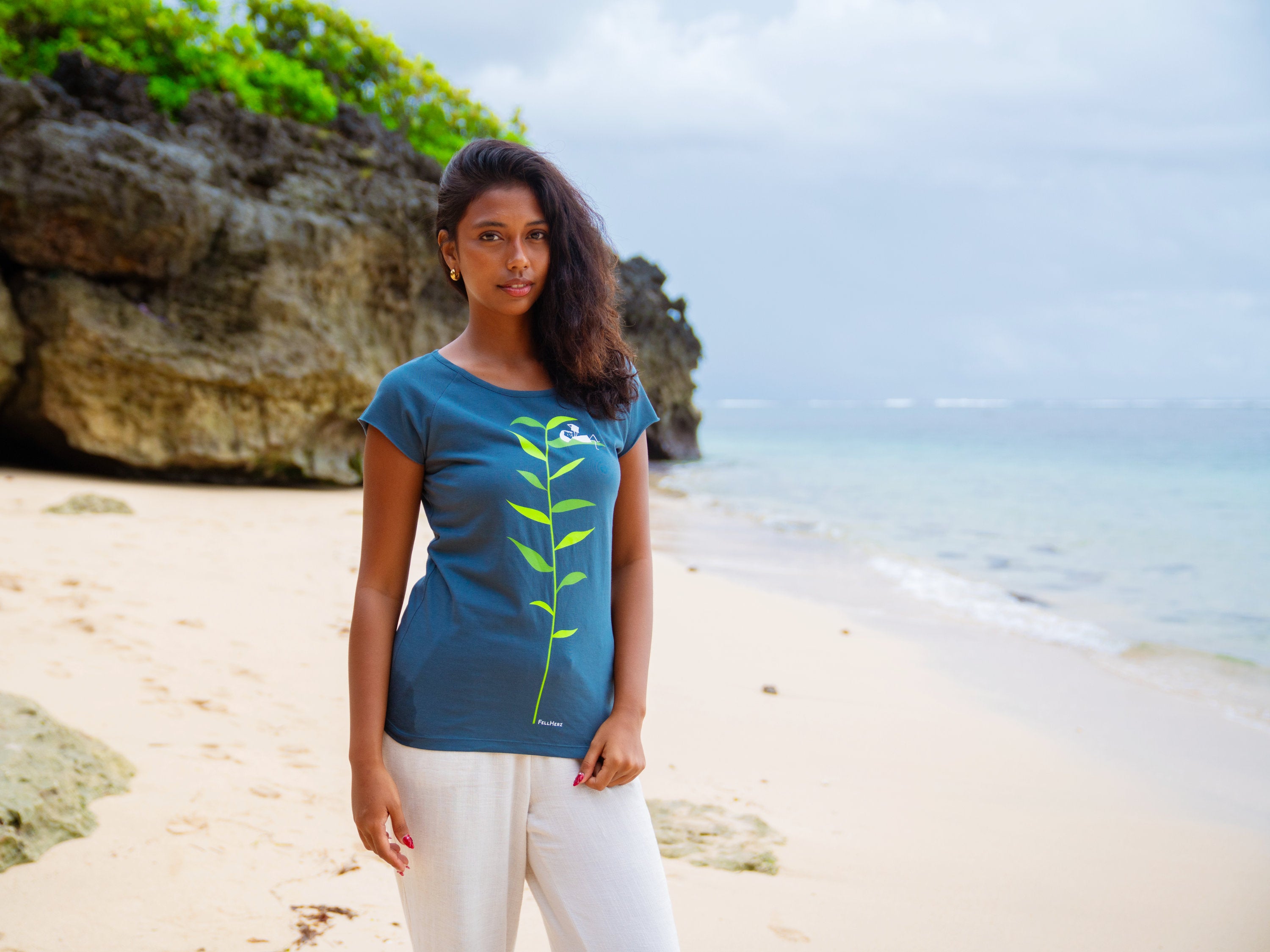 Ein weibliches Model steht am Strand und trägt ein graublaues FellHerz T-Shirt mit dem Aufdruck Booklover. Der Siebdruck stellt eine grüne Pflanze dar und auf einem Blatt liegt ein kleines Mädchen und liest aus einem Buch.