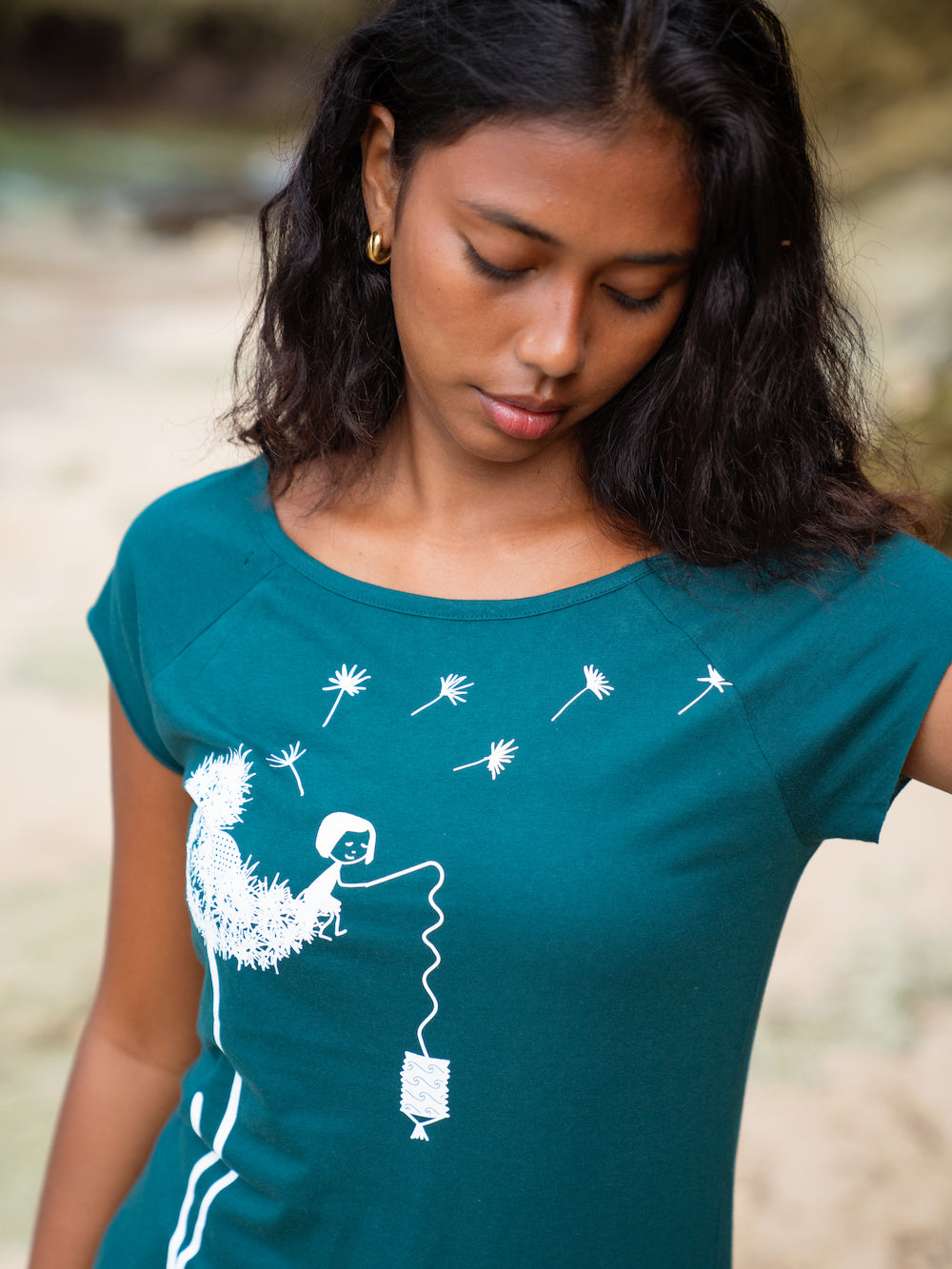 ein Modell steht am Strand und trägt das petrolfarbenes FellHerz Cap Sleeve Shirt Pusteblume. Das Motiv zeigt eine große weiße Pusteblume, auf der ein kleines Mädchen mit einer Laterne sitzt.