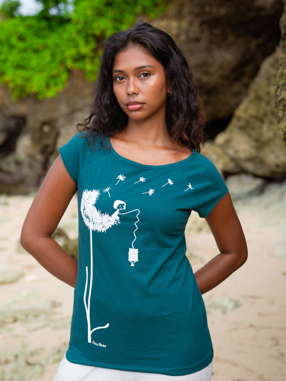 ein Modell steht am Strand und trägt das petrolfarbenes FellHerz Cap Sleeve Shirt Pusteblume. Das Motiv zeigt eine große weiße Pusteblume, auf der ein kleines Mädchen mit einer Laterne sitzt.
