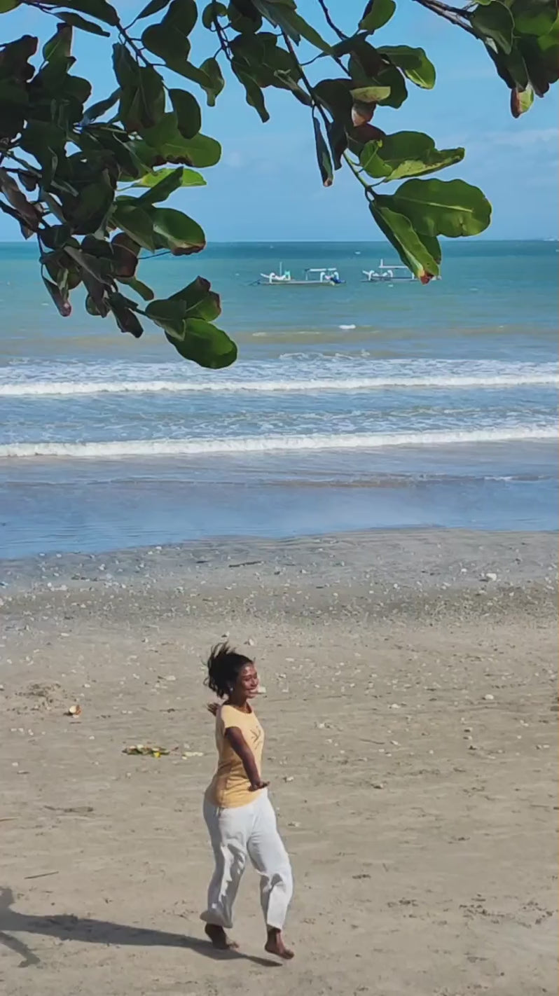 Video mit einem Model am Strand, das ein sandfarbenes Shirt mit auberginefarbenem Auftruck trägt. Die Grafik Lesepalme zeigt eine Pflanze, auf der ein kleines Mädchen sitzt, das in einem Buch liest.