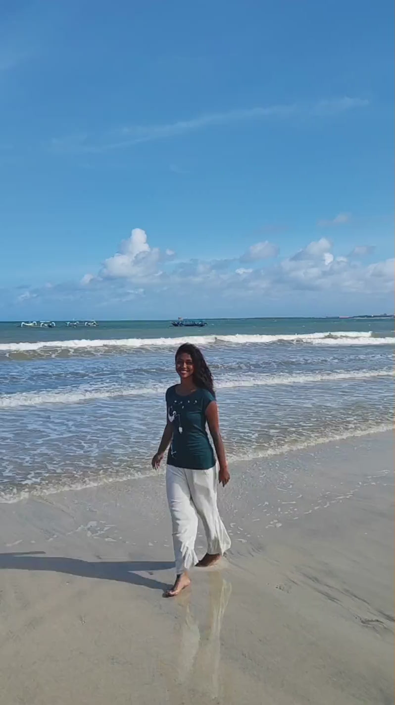 Ein Video zeigt ein Modell am Strand. Es trägt das petrolfarbene FellHerz Cap Sleeve Shirt Pusteblume. Das Motiv zeigt eine große weiße Pusteblume, auf der ein kleines Mädchen mit einer Laterne sitzt.
