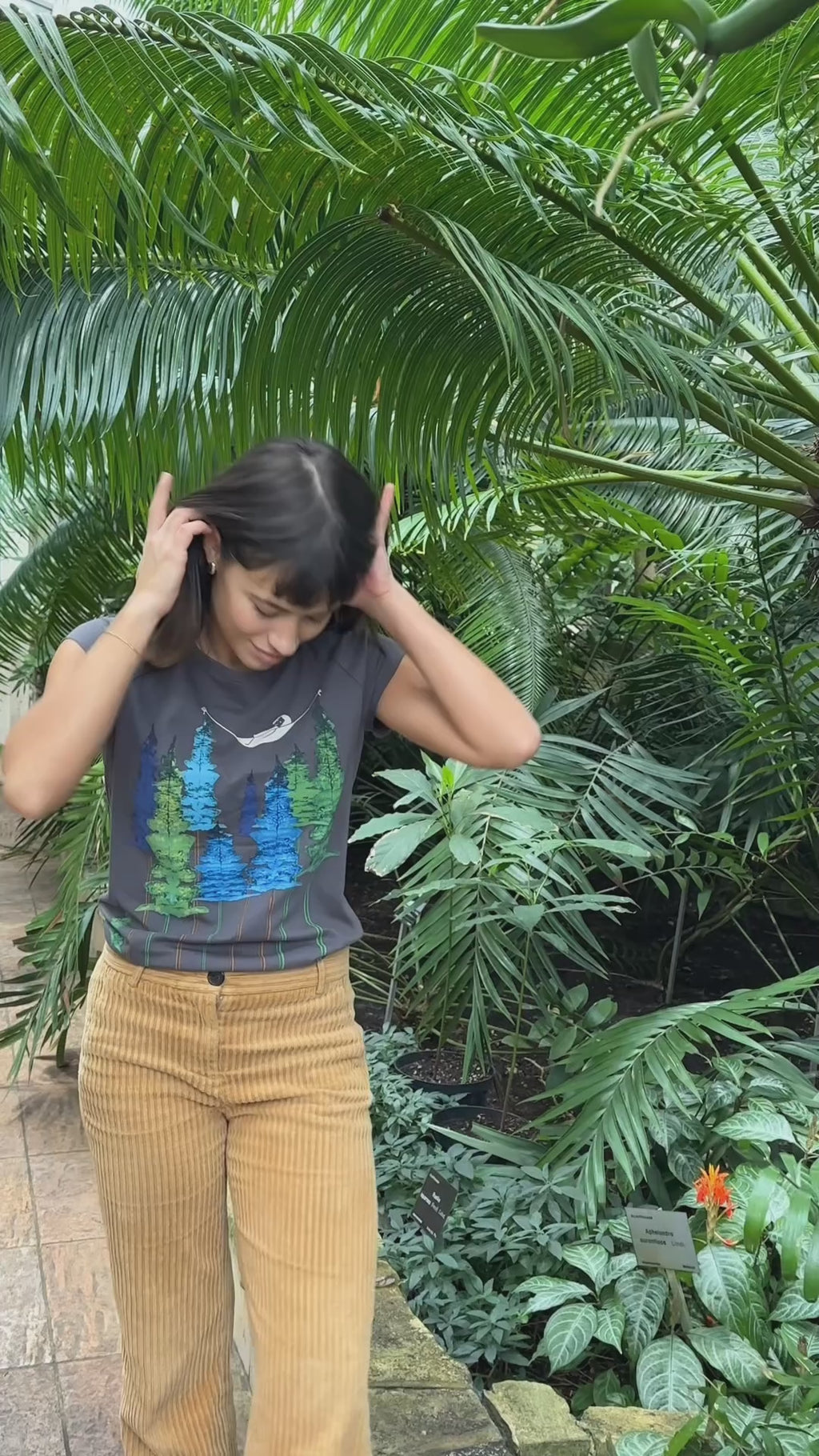 Video von Model in Botanischen Garten. Sie trägt ein Damen‑Cap‑Sleeve‑T‑Shirt in dunkelgrau, 100 % weiche GOTS‑zertifizierte Bio‑Baumwolle mit modernem, leicht tailliertem Schnitt und digitalem FellHerz‑Print mit einem kleinen Mädchen, dass in einer Hängematte, die zwischen Bäumen hängt liegt – nachhaltig, fair produziert, ideal für warme Tage oder als Lagen‑Basic.