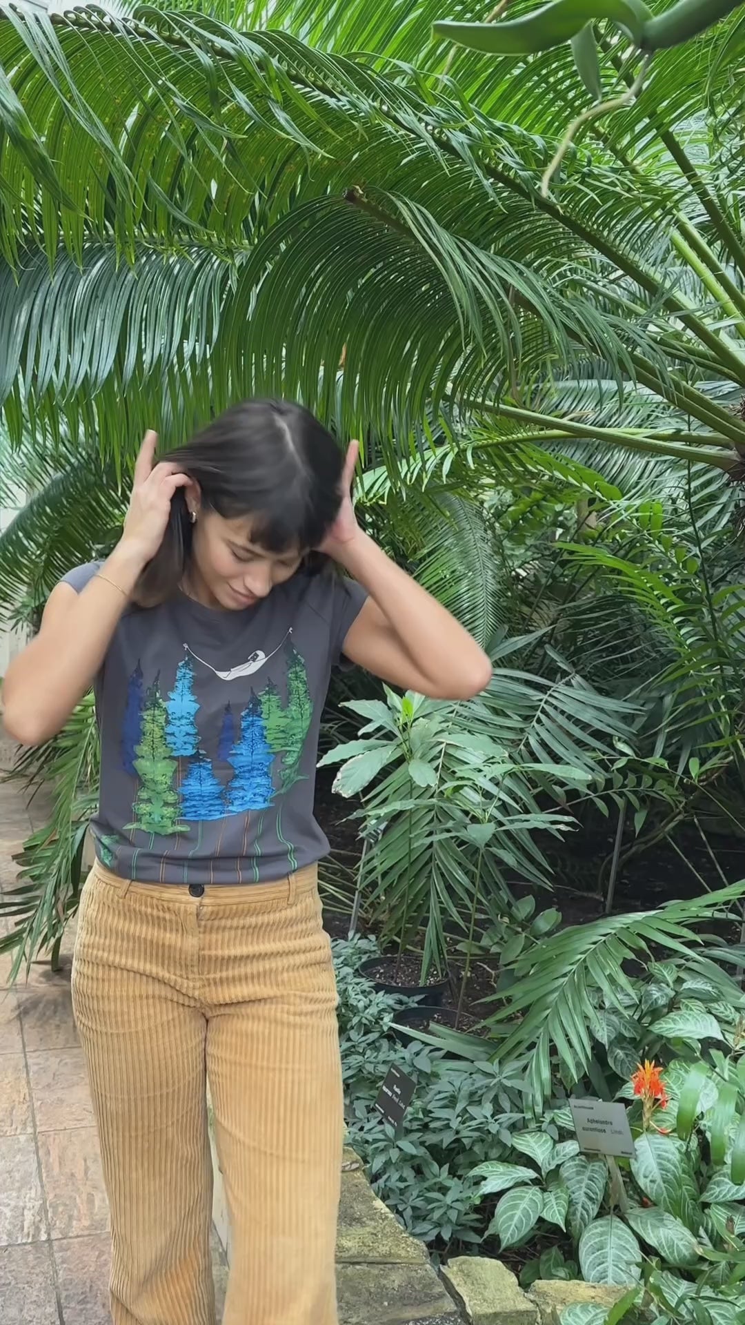 Video von Model in Botanischen Garten. Sie trägt ein Damen‑Cap‑Sleeve‑T‑Shirt in dunkelgrau, 100 % weiche GOTS‑zertifizierte Bio‑Baumwolle mit modernem, leicht tailliertem Schnitt und digitalem FellHerz‑Print mit einem kleinen Mädchen, dass in einer Hängematte, die zwischen Bäumen hängt liegt – nachhaltig, fair produziert, ideal für warme Tage oder als Lagen‑Basic.