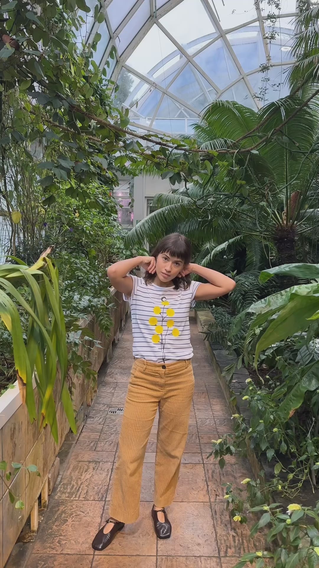 Video mit Model, sie trägt ein Weiß‑blau gestreiftes Damen‑T‑Shirt im Cap‑Sleeve‑Stil, leicht tailliert, mit weitem Rundhalsausschnitt aus weichem, GOTS‑zertifizierter Bio‑Baumwolle, nachhaltig und fair produziert. Auf der Vorderseite befindet sich ein Print: eine gelbe Blume, auf der sich ein kleines Mädchen ausruht. Das Shirt vermittelt ein entspanntes Urlaubs‑Feeling