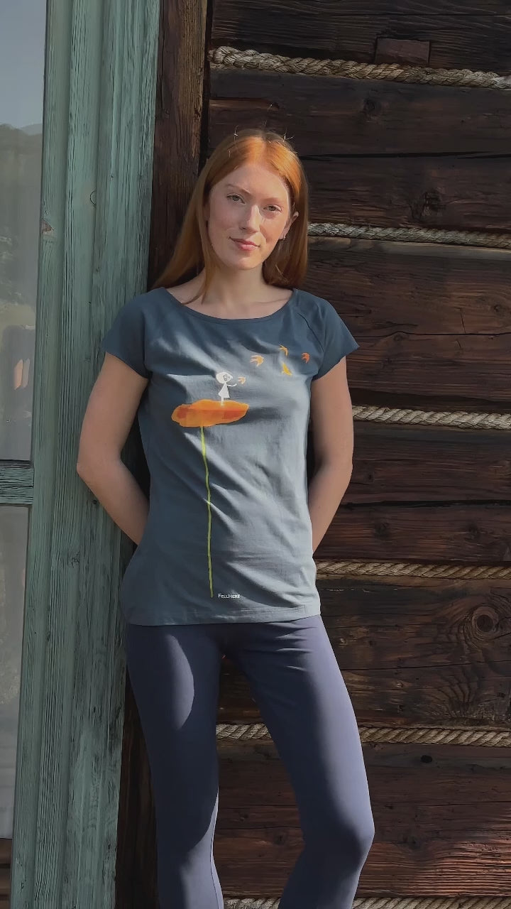 Video mit Model. Es trägt ein Mittel-blaues Damen T-Shirt aus Bio-Baumwolle, fair und vegan produziert. Raglan-Shirt mit Grafik-Print auf der Vorderseite: Ein Mädchen steht auf einer orangen Blume und es fliegen Vögel davon.