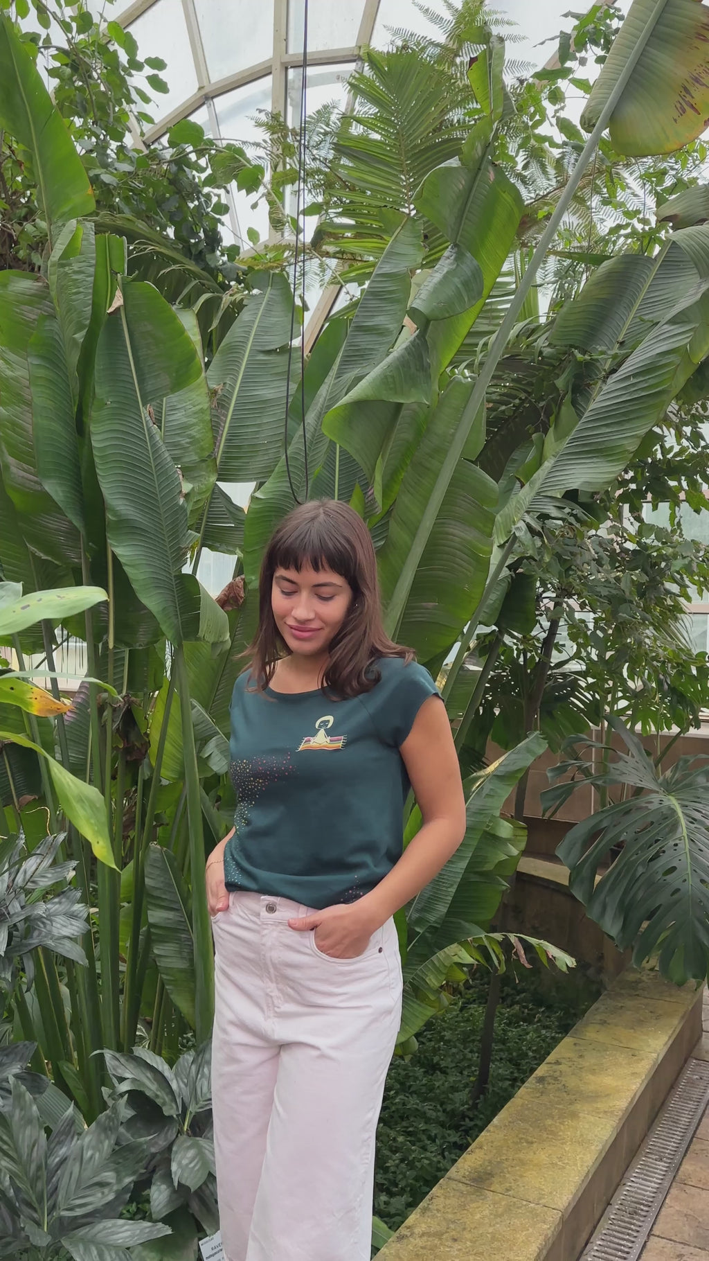 Video von Model in Botanischen Garten. Sie trägt ein Damen‑Cap‑Sleeve‑T‑Shirt in tiefem Deep Teal, 100 % weiche GOTS‑zertifizierte Bio‑Baumwolle mit modernem, leicht tailliertem Schnitt und digitalem FellHerz‑Print mit einem kleinen Mädchen, dass auf einem regenbogenfarbenen fliegenden Teppich sitzt und einen regenbogenen Schweif hinter sich her zieht – nachhaltig, fair produziert, ideal für warme Tage oder als Lagen‑Basic.