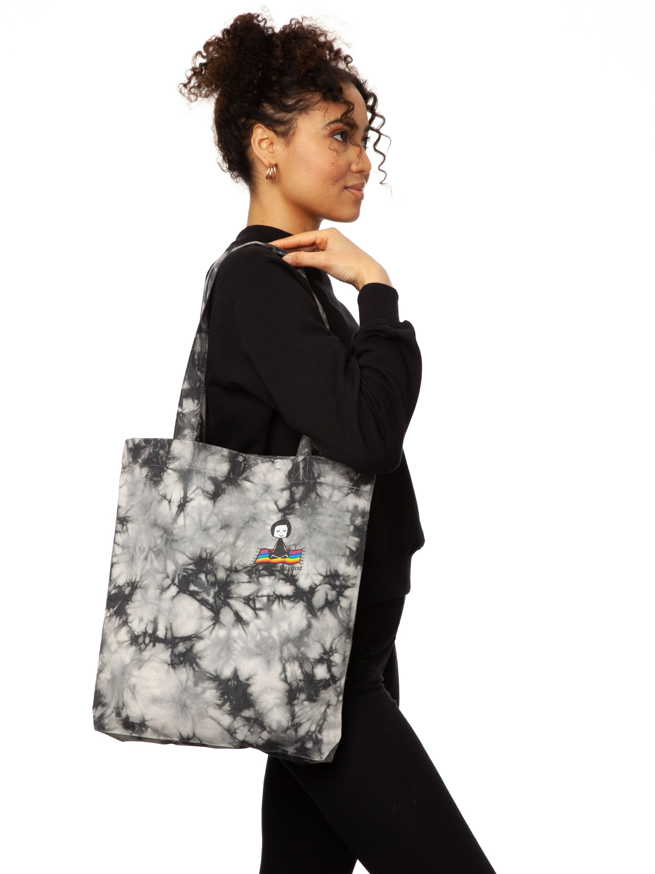 Schwarz weiße Batik‑Tote‑Bag ‘Ommm’ in handgefärbtem Tie‑Dye‑Muster, ca. 37 × 39 cm, aus 300 g/m² Canvas (80 % recycelte Baumwolle, 20 % recycelter Polyester), mit robust verstärkten Kanten, extra Volumen durch Bodennaht und digitalem Ommm‑Print, auf dem ein Mädchen auf einem regenbogenfarbenen Teppich fliegt – fair produziert (Fair Wear), vorgewaschen, waschbar bei 30 °C