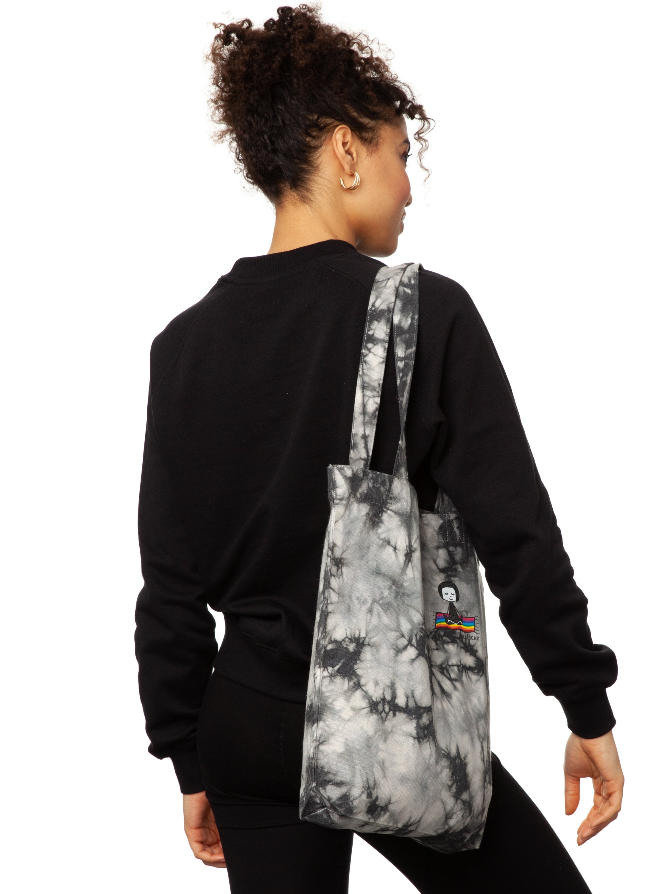 Schwarz weiße Batik‑Tote‑Bag ‘Ommm’ in handgefärbtem Tie‑Dye‑Muster, ca. 37 × 39 cm, aus 300 g/m² Canvas (80 % recycelte Baumwolle, 20 % recycelter Polyester), mit robust verstärkten Kanten, extra Volumen durch Bodennaht und digitalem Ommm‑Print, auf dem ein Mädchen auf einem regenbogenfarbenen Teppich fliegt – fair produziert (Fair Wear), vorgewaschen, waschbar bei 30 °C
