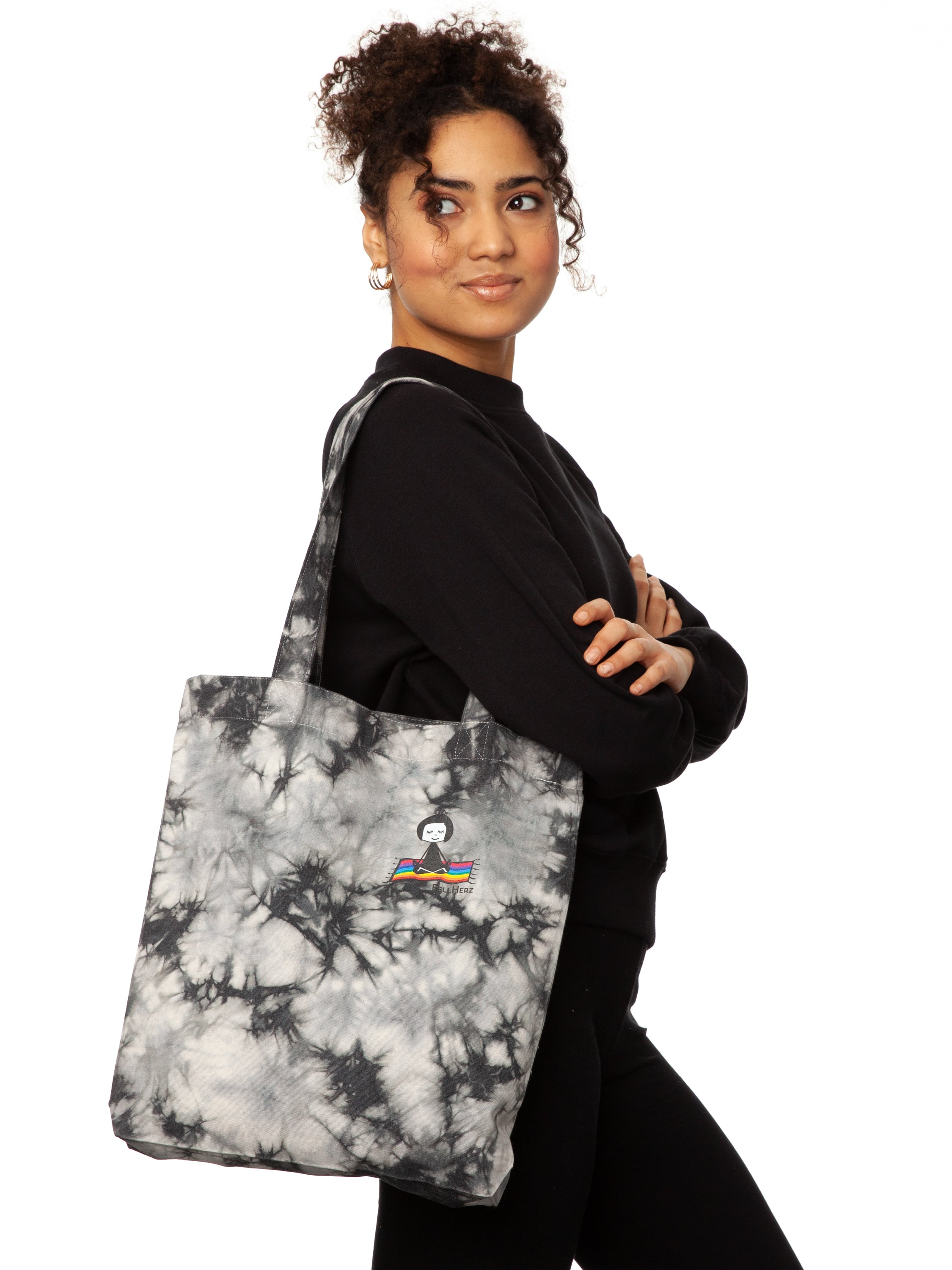 Schwarz weiße Batik‑Tote‑Bag ‘Ommm’ in handgefärbtem Tie‑Dye‑Muster, ca. 37 × 39 cm, aus 300 g/m² Canvas (80 % recycelte Baumwolle, 20 % recycelter Polyester), mit robust verstärkten Kanten, extra Volumen durch Bodennaht und digitalem Ommm‑Print, auf dem ein Mädchen auf einem regenbogenfarbenen Teppich fliegt – fair produziert (Fair Wear), vorgewaschen, waschbar bei 30 °C