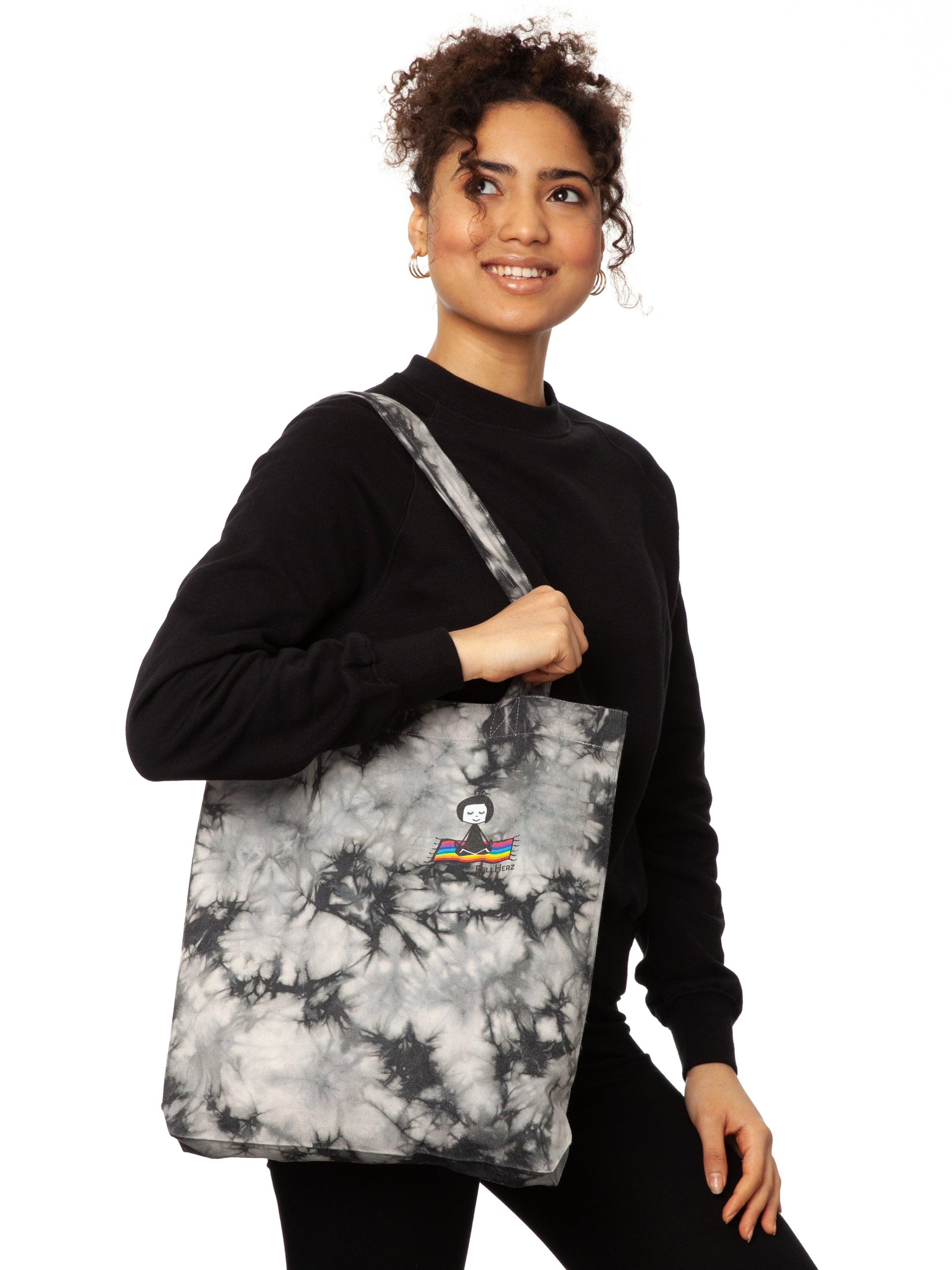 Schwarz weiße Batik‑Tote‑Bag ‘Ommm’ in handgefärbtem Tie‑Dye‑Muster, ca. 37 × 39 cm, aus 300 g/m² Canvas (80 % recycelte Baumwolle, 20 % recycelter Polyester), mit robust verstärkten Kanten, extra Volumen durch Bodennaht und digitalem Ommm‑Print, auf dem ein Mädchen auf einem regenbogenfarbenen Teppich fliegt – fair produziert (Fair Wear), vorgewaschen, waschbar bei 30 °C