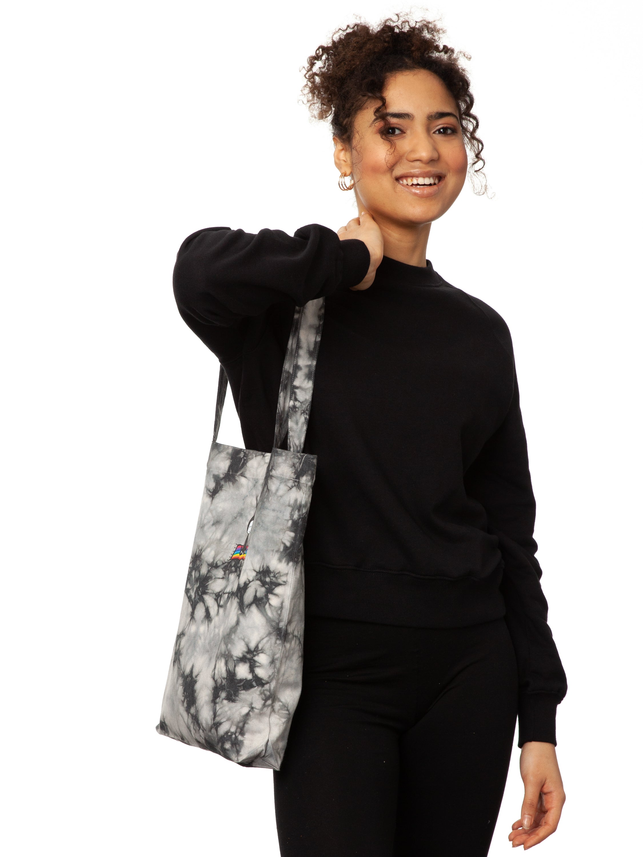 Schwarz weiße Batik‑Tote‑Bag ‘Ommm’ in handgefärbtem Tie‑Dye‑Muster, ca. 37 × 39 cm, aus 300 g/m² Canvas (80 % recycelte Baumwolle, 20 % recycelter Polyester), mit robust verstärkten Kanten, extra Volumen durch Bodennaht und digitalem Ommm‑Print, auf dem ein Mädchen auf einem regenbogenfarbenen Teppich fliegt – fair produziert (Fair Wear), vorgewaschen, waschbar bei 30 °C